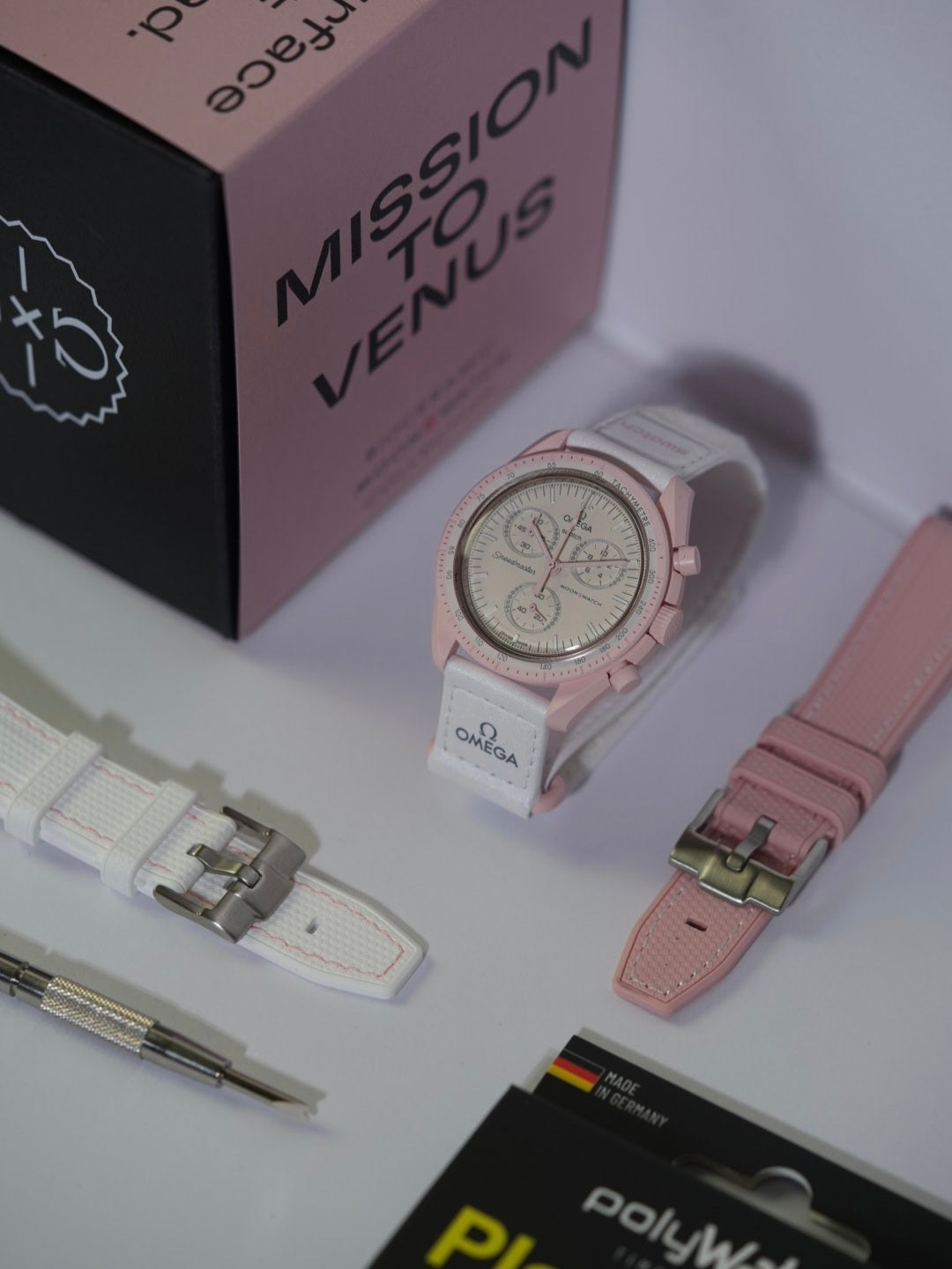 Swatch x Omega MoonSwatch Mission to Venus watch displayed on a stand, featuring a soft pink bioceramic case, light pink dial, and white strap with pink stitching. The bundle includes additional strap options, including a textured white strap with pink stitching and a pastel pink leather-style strap. A spring bar tool and a PolyWatch Plastic Polish kit are also included, with the original 'Mission to Venus' packaging box in the background