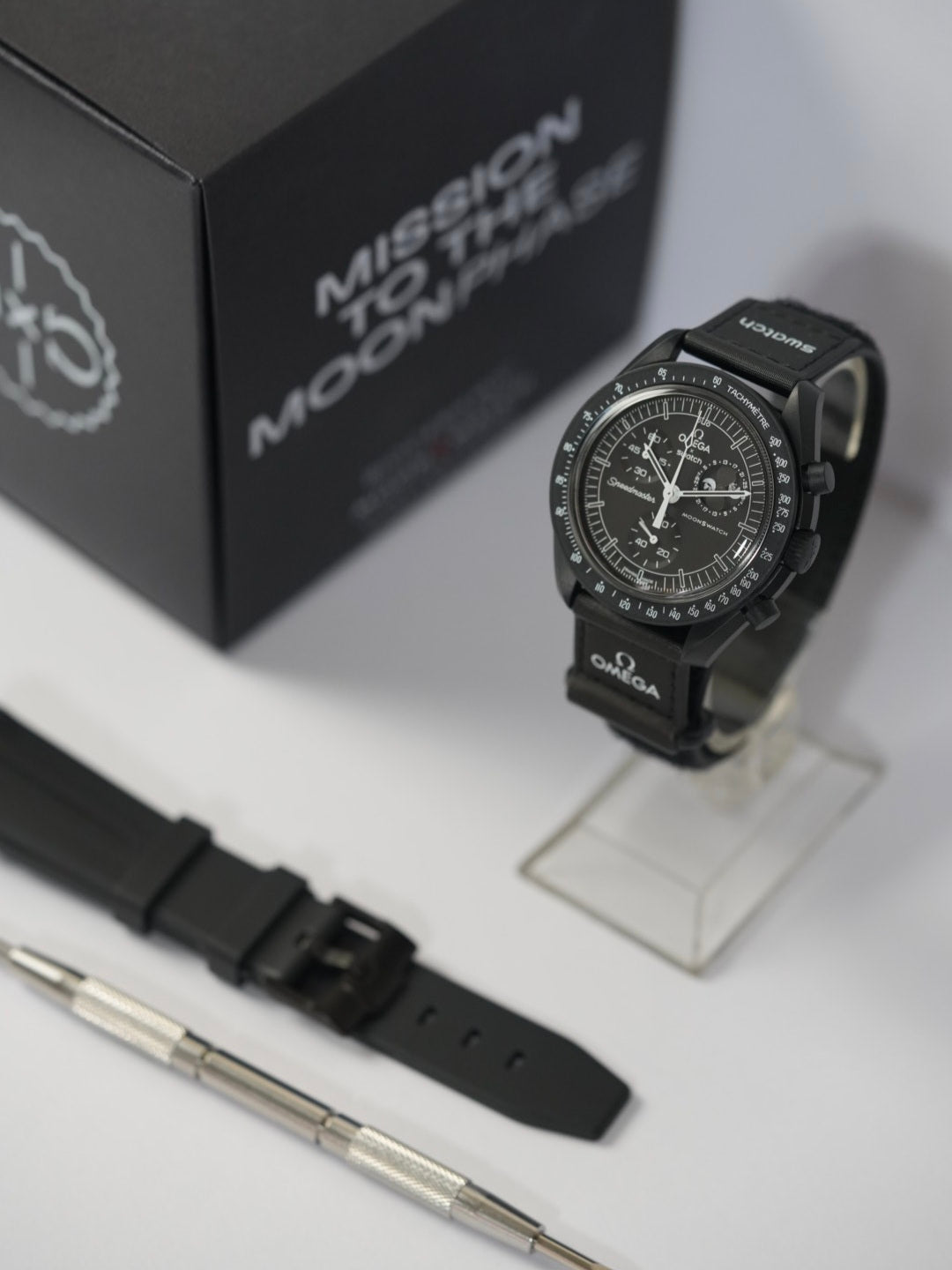 Swatch x Omega MoonSwatch Mission to the MoonPhase watch displayed on a stand, with its black strap featuring Omega and Swatch branding. A black watch strap, a spring bar tool, and the original packaging box labeled 'Mission to the MoonPhase' are also arranged on a white surface