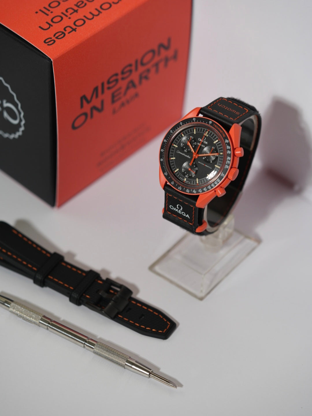 Swatch x Omega MoonSwatch Mission on Earth Lava bundle featuring the watch, an additional black strap with orange stitching, and a spring bar tool, displayed with its packaging.