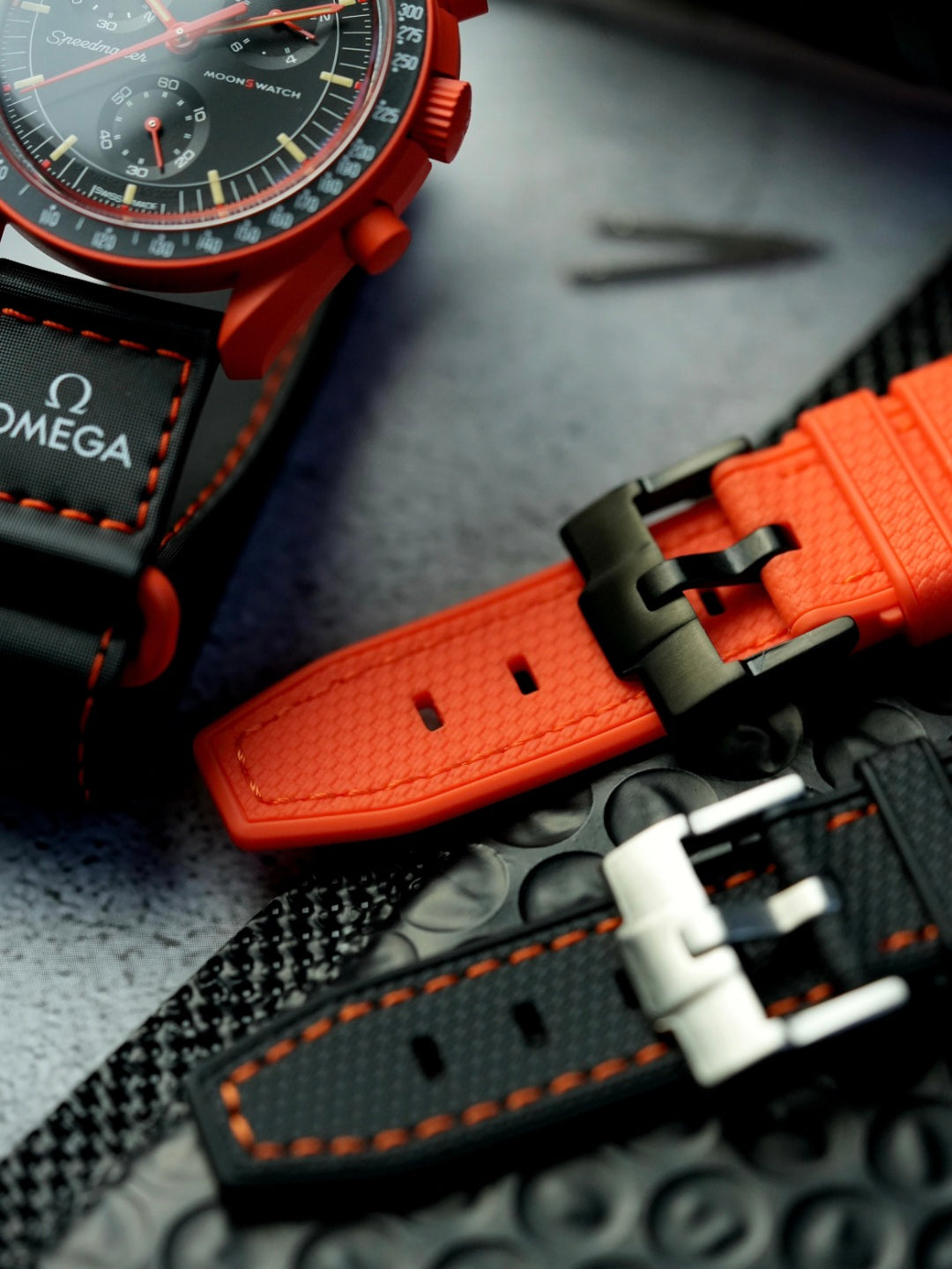 Swatch x Omega MoonSwatch Mission on Earth Lava watch with two premium rubber straps—one in bold orange with a black buckle and one in black with orange stitching—displayed on a textured surface.