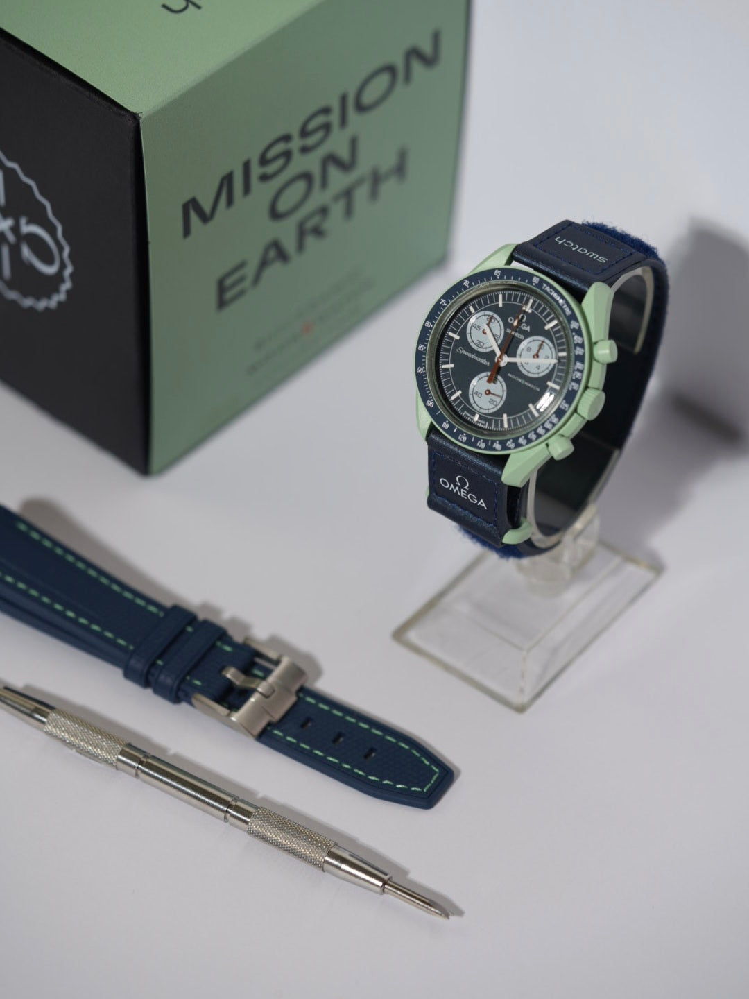 Swatch x Omega MoonSwatch Mission on Earth watch bundle featuring the timepiece, a navy blue strap with green stitching, and a spring bar tool, displayed with its packaging.