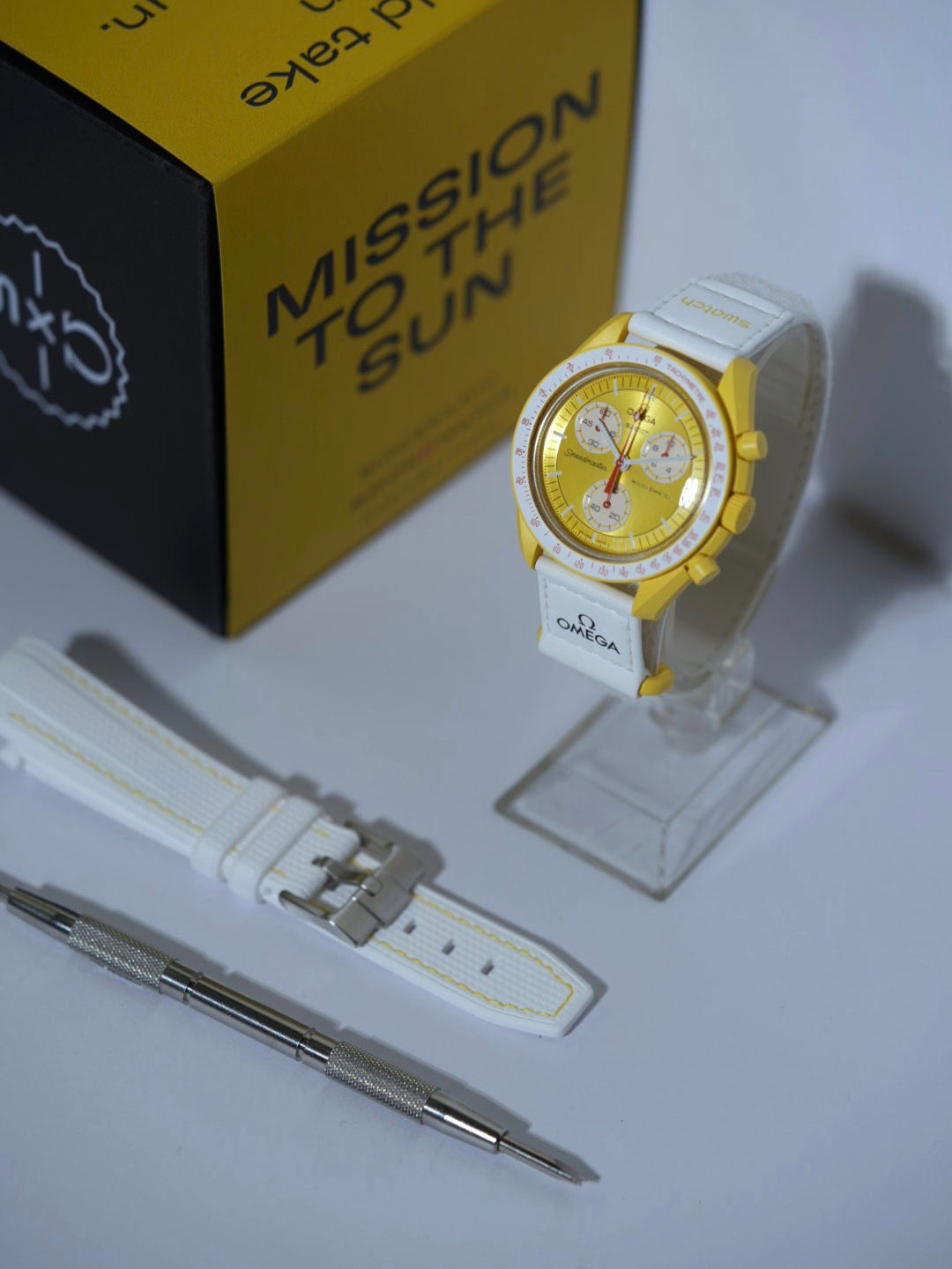 Swatch x Omega MoonSwatch Mission to the Sun bundle featuring the Sun watch, a white rubber strap with yellow stitching, and a spring bar tool, displayed with its packaging.