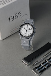 Gray 1965 MoonSwatch watch with white face and black accents on a gray surface, with '1965' text in the background.