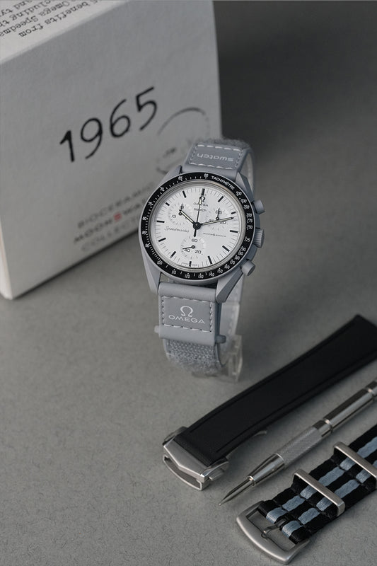 Gray 1965 MoonSwatch watch with white face and black accents on a gray surface, with '1965' text in the background.