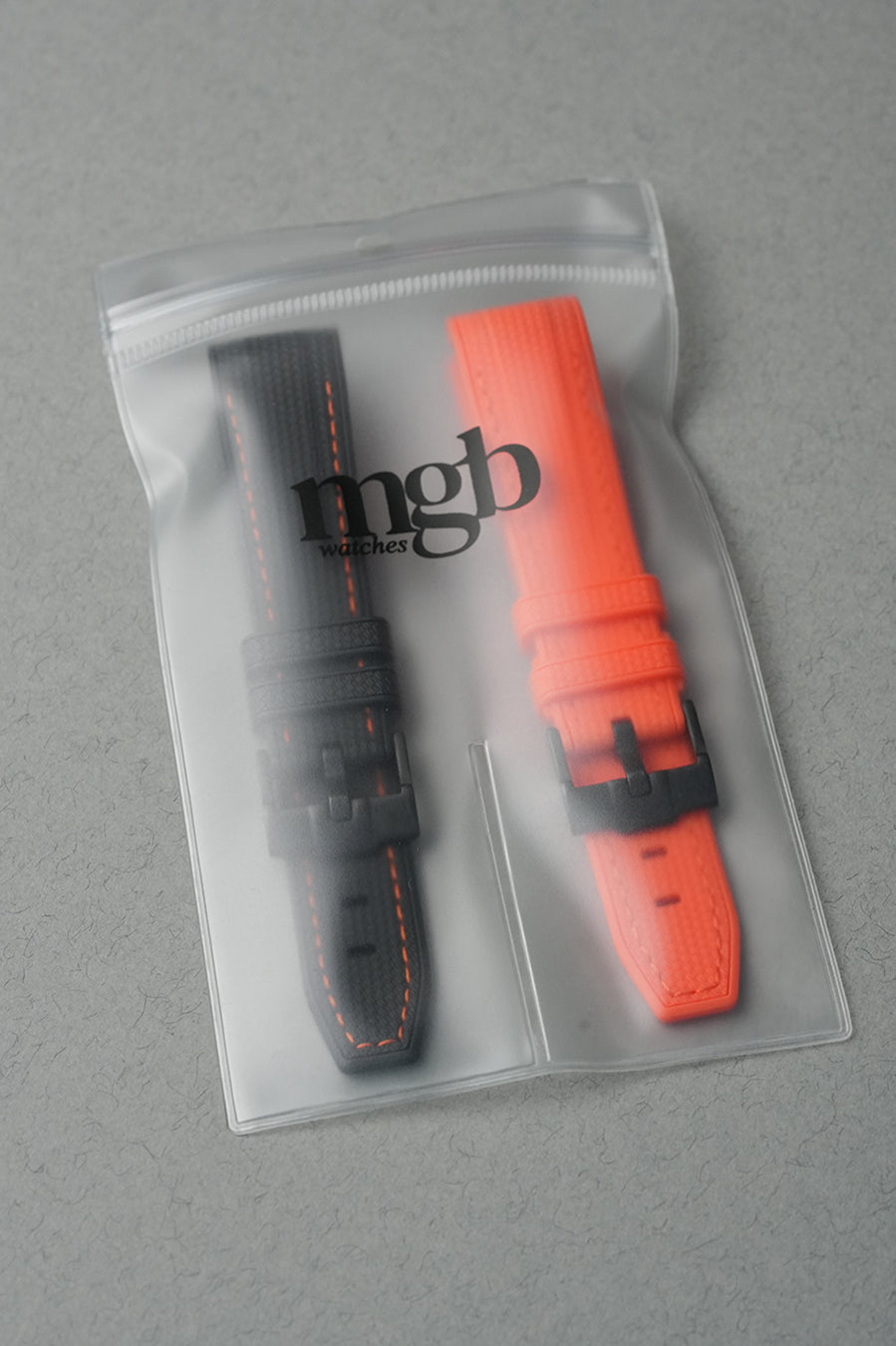 Two watch straps, one black and one orange, in a clear packaging bag on a gray background.