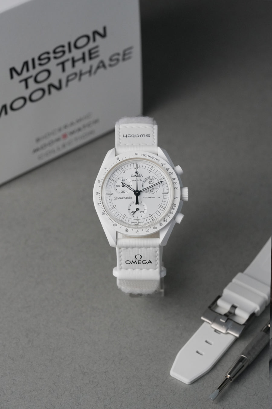 Omega MoonSwatch with white strap on a gray surface, branded packaging in the background.
