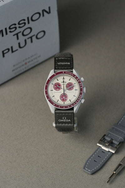Omega watch with black leather strap on a gray surface, with 'Mission to Pluto' box in the background.