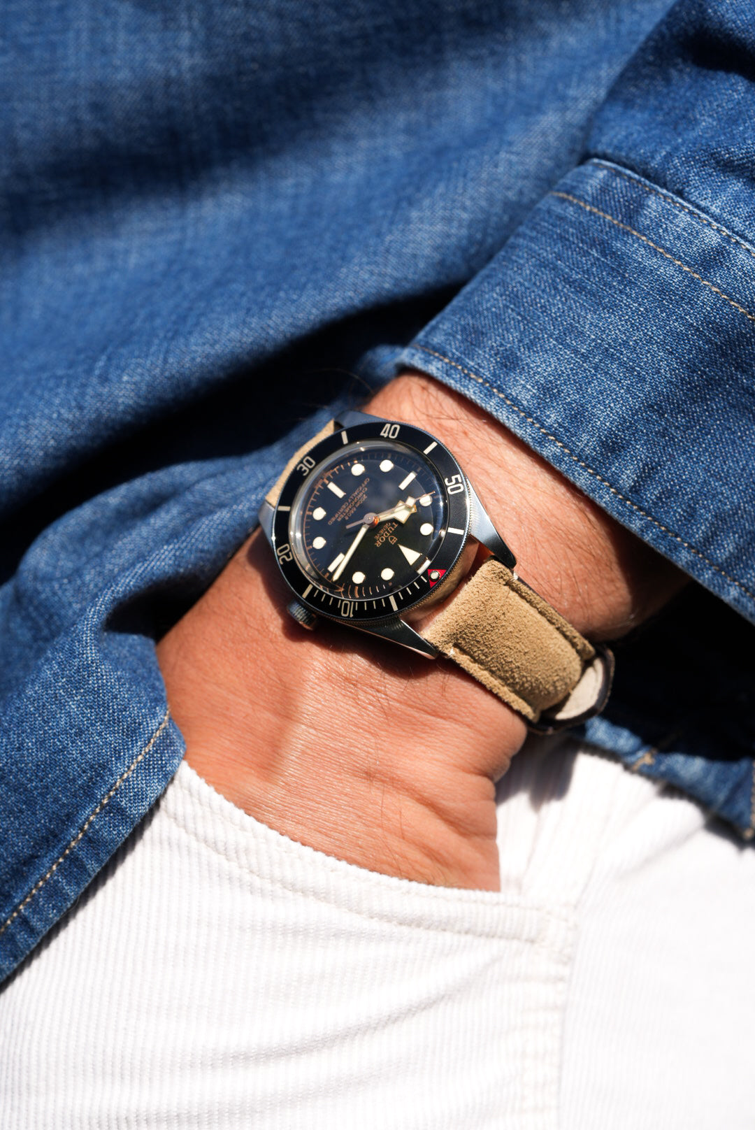 Close-up of Tudor Black Bay 58 watch on beige suede strap, styled casually with denim shirt and white trousers