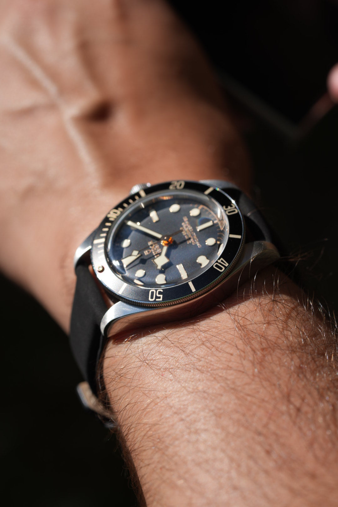 Close-up of a Tudor Black Bay 58 watch on a black nubuck strap, showing brushed steel case and black bezel in sunlight.