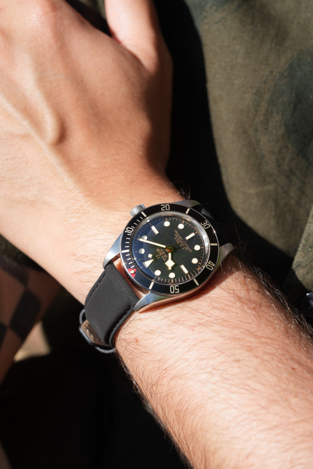 Tudor Black Bay 58 on wrist with black nubuck strap, worn casually with tattooed arm and olive shirt in background.