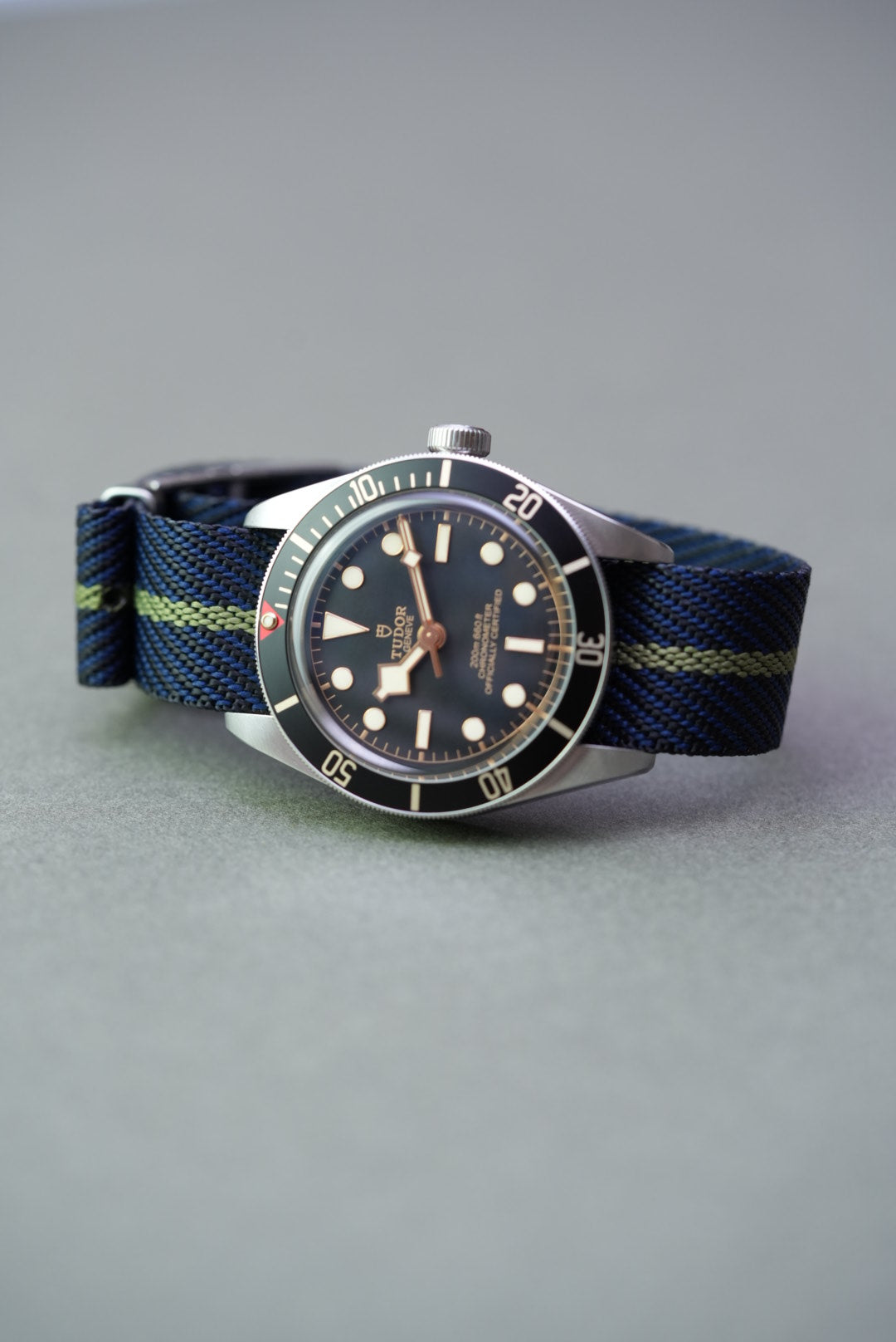 Tudor Black Bay on a navy woven strap with green centre stripe, laid flat on grey background.