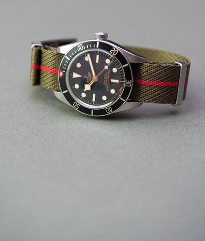 Tudor Black Bay watch on an olive green woven nylon strap with bold red centre stripe, shown from front angle.