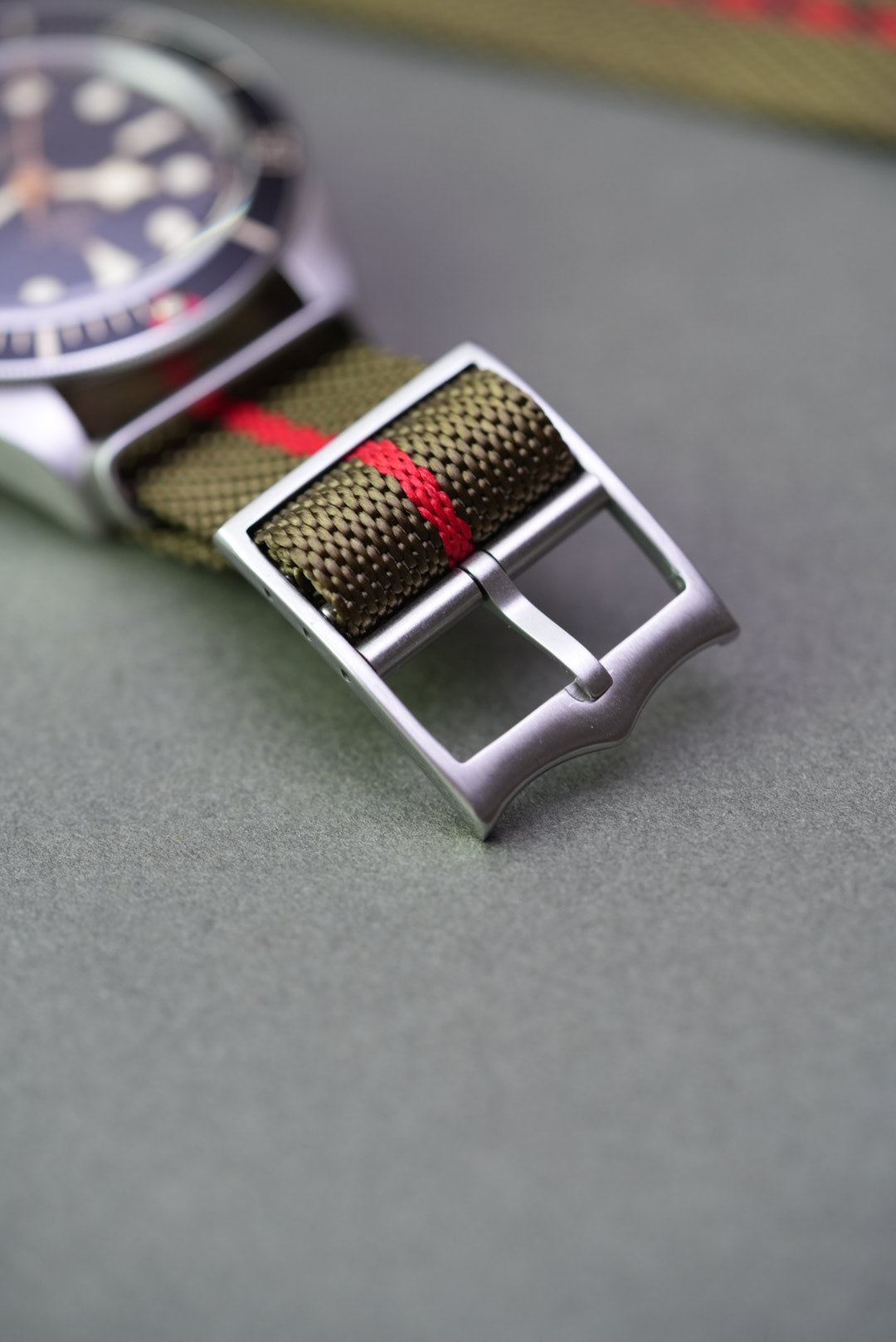Close-up of brushed steel buckle on an olive green woven nylon strap with red centre stripe, fitted to a Tudor Black Bay watch.