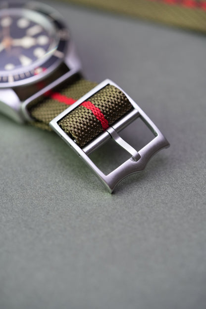 Close-up of brushed steel buckle on an olive green woven nylon strap with red centre stripe, fitted to a Tudor Black Bay watch.